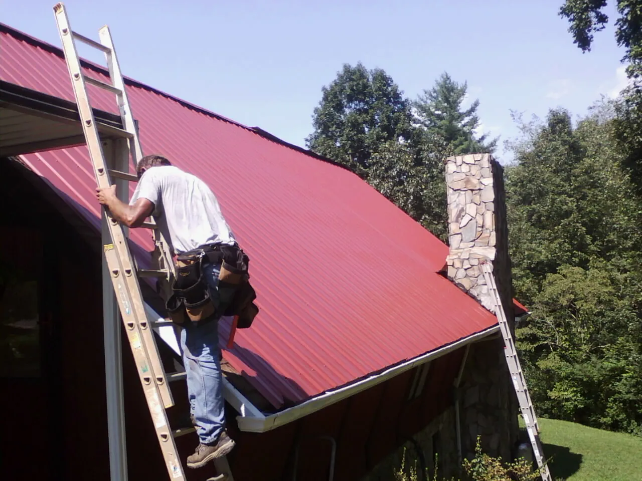 Roofer on a ladder beside a red metal roof and stone chimney — Soffit & Fascia Repair by Roofing Brothers in Greencastle